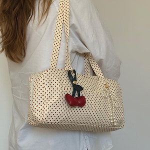 May include: A rectangular cream-colored shoulder bag featuring a small red polka dot pattern. The bag includes two shoulder straps, a red cherry-shaped charm with green leaves, and a dangling white faux pearl beaded accessory attached to the side.