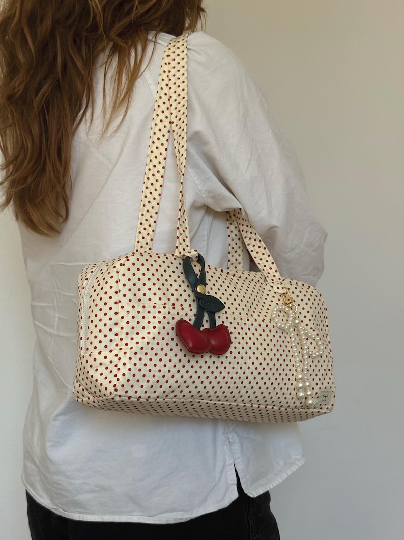 May include: A rectangular cream-colored shoulder bag featuring a small red polka dot pattern. The bag includes two shoulder straps, a red cherry-shaped charm with green leaves, and a dangling white faux pearl beaded accessory attached to the side.