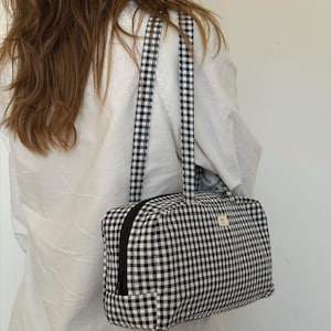 May include: A rectangular shoulder bag featuring a black and white gingham check pattern. The bag has two long fabric straps, a black zipper closure along the top, and a small rectangular label on the front with black text.