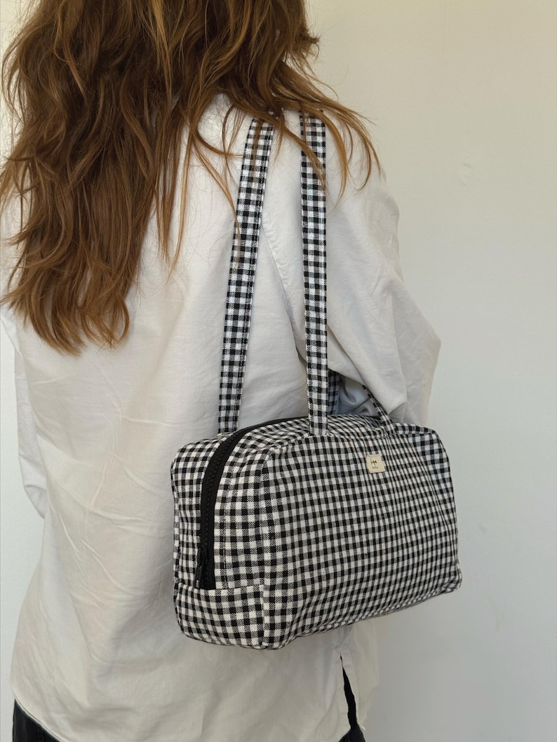 May include: A rectangular shoulder bag featuring a black and white gingham check pattern. The bag has two long fabric straps, a black zipper closure along the top, and a small rectangular label on the front with black text.