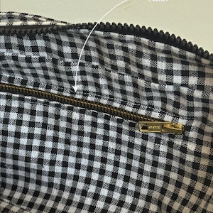 May include: The interior of a bag featuring a black and white gingham check pattern. A horizontal brass-toned metal zipper is integrated into the fabric, labeled with the text ZIP INTERIOR POCKET, providing access to a secure storage compartment.