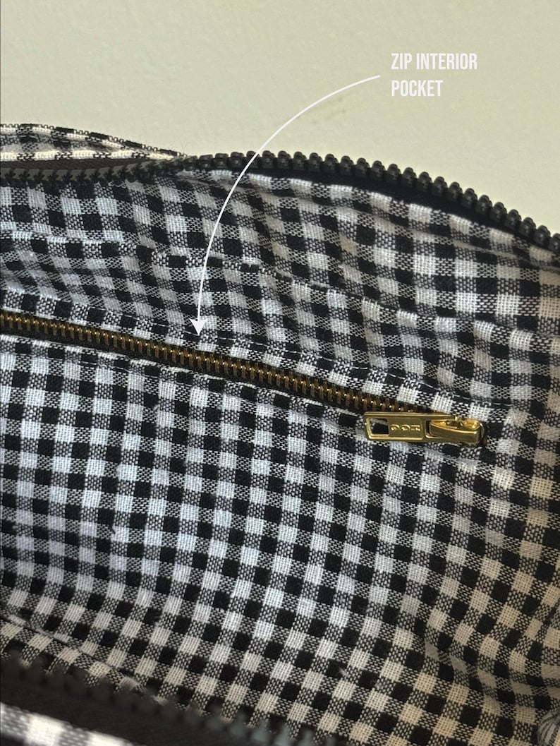 May include: The interior of a bag featuring a black and white gingham check pattern. A horizontal brass-toned metal zipper is integrated into the fabric, labeled with the text ZIP INTERIOR POCKET, providing access to a secure storage compartment.