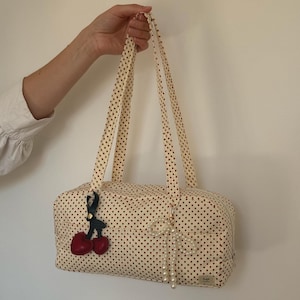 May include: A rectangular fabric bag with a cream base and small red polka dots, featuring two long shoulder straps. The bag is decorated with a red cherry charm on a black strap and a white pearl-like beaded bow charm hanging from the front.