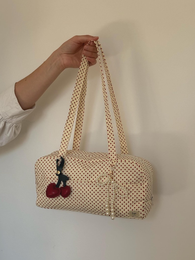 May include: A rectangular fabric bag with a cream base and small red polka dots, featuring two long shoulder straps. The bag is decorated with a red cherry charm on a black strap and a white pearl-like beaded bow charm hanging from the front.