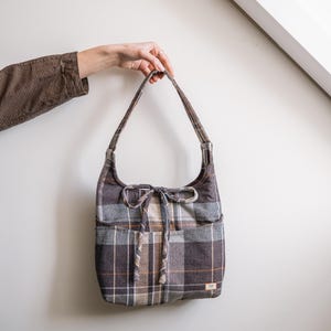 Op de afbeelding: Een geruite schoudertas in bruin, grijs en beige, met een trekkoordsluiting en twee buitenzakken. De tas wordt door een hand omhoog gehouden. Het ontwerp van de tas heeft een herhalend patroon van elkaar kruisende lijnen.