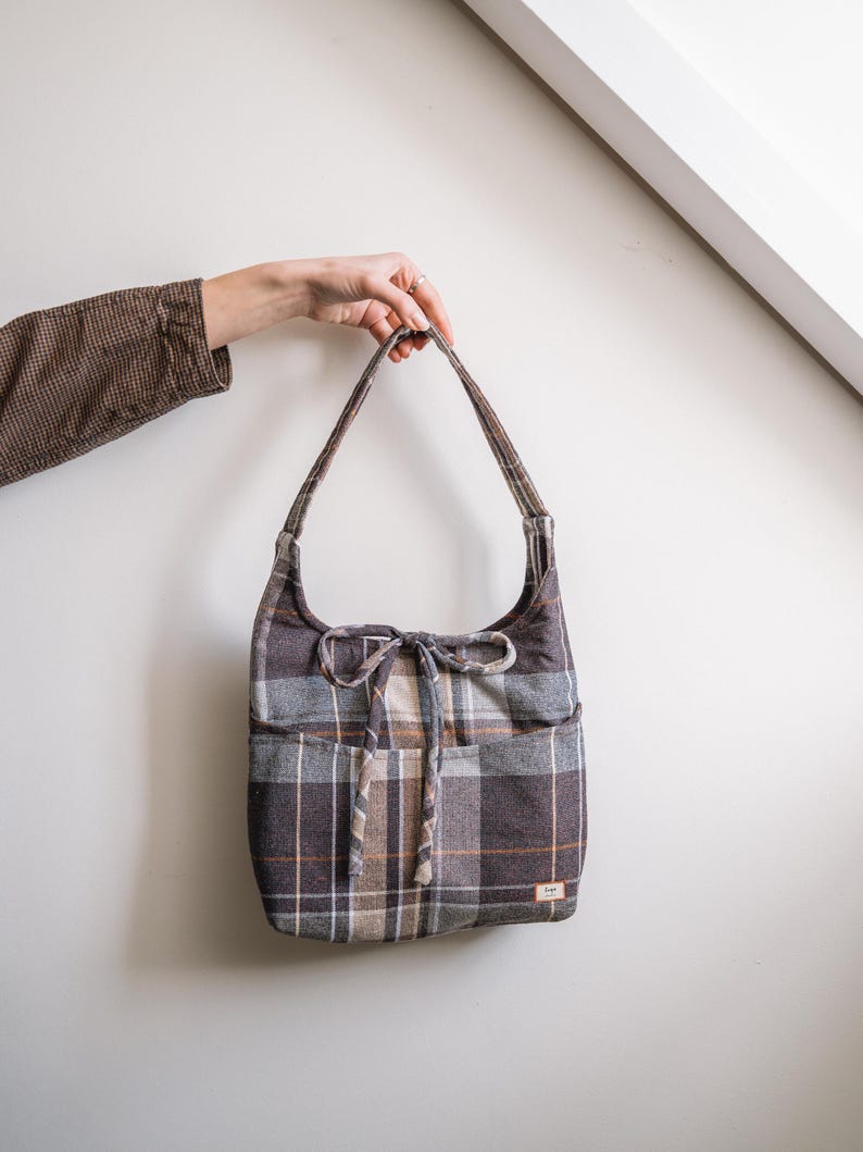 Op de afbeelding: Een geruite schoudertas in bruin, grijs en beige, met een trekkoordsluiting en twee buitenzakken. De tas wordt door een hand omhoog gehouden. Het ontwerp van de tas heeft een herhalend patroon van elkaar kruisende lijnen.