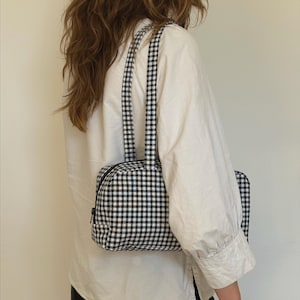 May include: A rectangular shoulder bag featuring a black and white gingham check pattern. The bag has a top zipper closure with a black pull tab and two matching fabric shoulder straps. The overall design is structured with a flat base and sides.