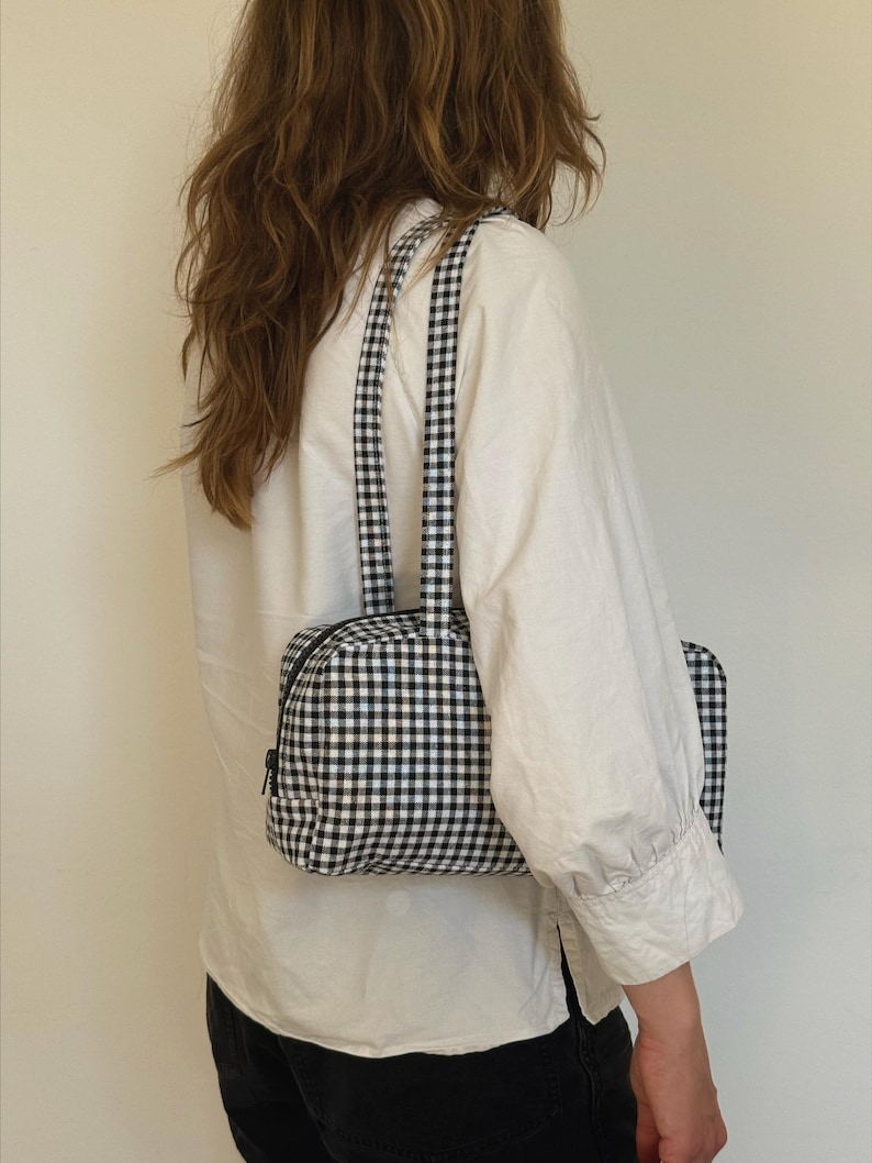 May include: A rectangular shoulder bag featuring a black and white gingham check pattern. The bag has a top zipper closure with a black pull tab and two matching fabric shoulder straps. The overall design is structured with a flat base and sides.