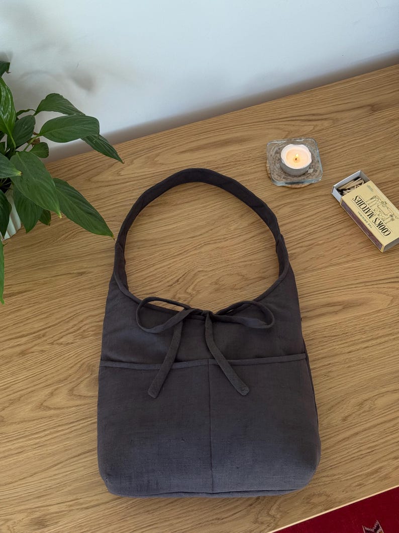 Op de afbeelding: Een donkergrijze schoudertas met een gebogen handvat en een strikdetail. De tas heeft twee voorzakken en is gemaakt van een zachte, getextureerde stof. De tas ligt op een houten oppervlak.