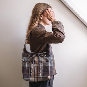 Op de afbeelding: Een geruite schoudertas in bruin, grijs en beige. De tas heeft een afgeronde vorm, een trekkoordsluiting en twee voorzakken. De riem is gemaakt van dezelfde geruite stof. Een klein label is op de voorkant genaaid.