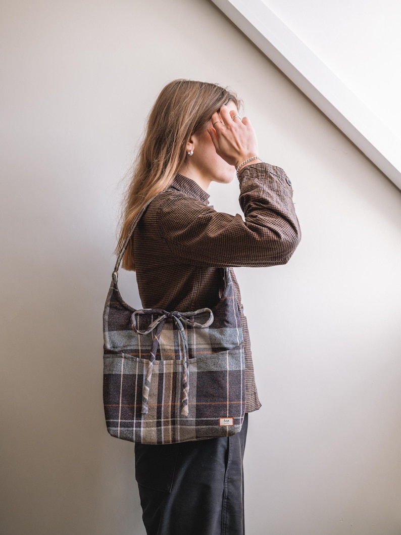 Op de afbeelding: Een geruite schoudertas in bruin, grijs en beige. De tas heeft een afgeronde vorm, een trekkoordsluiting en twee voorzakken. De riem is gemaakt van dezelfde geruite stof. Een klein label is op de voorkant genaaid.