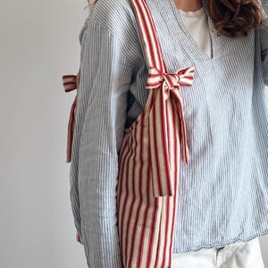 Op de afbeelding: Een rood-cr&egrave;me gestreepte tote bag met bijpassende bandjes en strikdetails. De tas heeft een rechthoekige vorm met een klein vakje aan de voorkant. De bandjes zijn bovenaan met strikken vastgemaakt. De tas is gemaakt van een zachte stof.