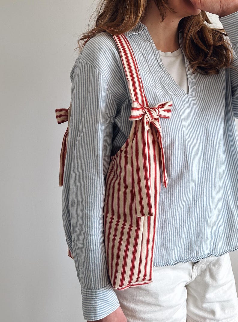 Op de afbeelding: Een rood-cr&egrave;me gestreepte tote bag met bijpassende bandjes en strikdetails. De tas heeft een rechthoekige vorm met een klein vakje aan de voorkant. De bandjes zijn bovenaan met strikken vastgemaakt. De tas is gemaakt van een zachte stof.