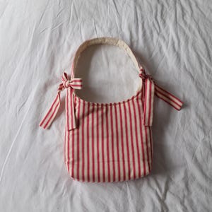 Op de afbeelding: Een rood-wit gestreepte schoudertas met een afgeronde vorm. De tas heeft een handvat en is aan elke kant met strikken vastgebonden. De tas is gemaakt van stof en staat tegen een witte achtergrond.