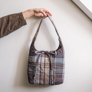 Op de afbeelding: Een gewatteerde tote bag met een ruitpatroon in bruin-, grijs- en oranjetinten. De tas heeft lange banden en een strikdetail aan de bovenkant. Een klein label is aan de voorkant van de tas bevestigd.