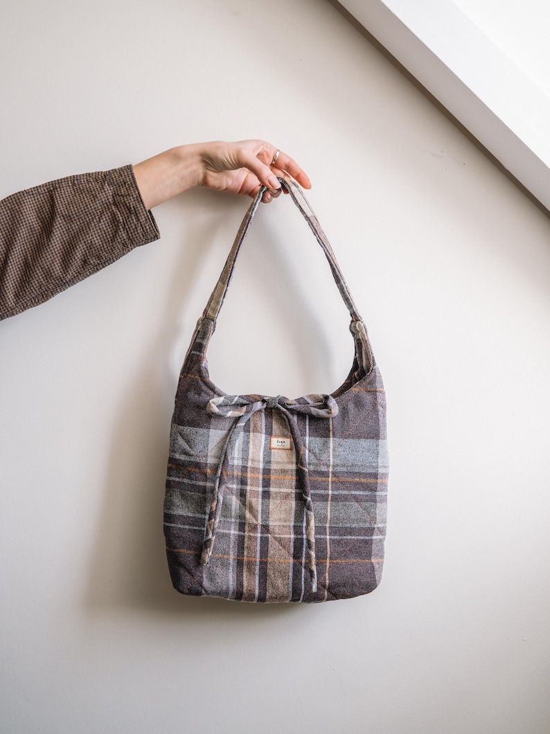 Op de afbeelding: Een gewatteerde tote bag met een ruitpatroon in bruin-, grijs- en oranjetinten. De tas heeft lange banden en een strikdetail aan de bovenkant. Een klein label is aan de voorkant van de tas bevestigd.