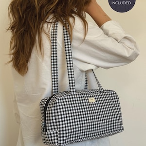 May include: A rectangular shoulder bag featuring a black and white gingham check pattern, two matching fabric straps, and a black zipper closure. A small rectangular label is attached to the front. A circular badge states VIDEO TUTORIAL INCLUDED.