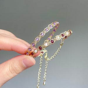May include: Two gold bracelets with purple and red gemstones. The bracelets are linked together with a gold chain.