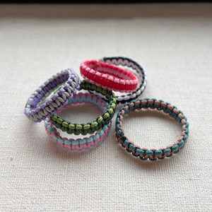May include: Five colourful braided rings made of different coloured threads. The rings are stacked on top of each other and are made of purple, green, pink, red and brown thread.