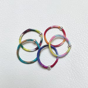 May include: Five colorful hair ties arranged on a white surface. Each tie is a different color, including red, orange, yellow, green, blue, and purple. Each tie has a small silver bead. These hair accessories are perfect for adding a pop of color to any hairstyle.