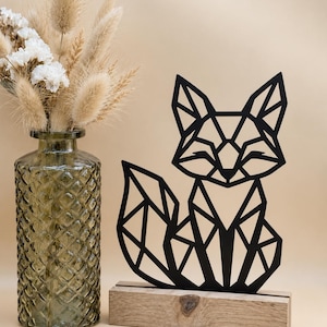 May include: A black geometric fox silhouette crafted from thin material, standing upright in a rectangular light brown wooden base. Beside it sits a tall, clear green glass vase with a diamond-textured pattern, filled with dried beige grasses.