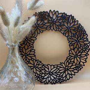 Puede incluir: Una corona floral negra con un diseño delicado e intrincado. La corona está hecha de un material negro delgado y tiene una forma circular. La corona está sentada sobre una superficie marrón claro.