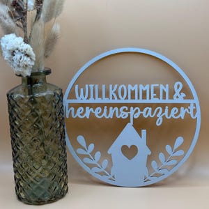 Könnte beinhalten: Ein hellblaues Holzschild mit dem Text "Willkommen & hereinspaziert" in einem Kreis mit einem Haus und Blättern. Das Schild hängt an einer Wand.