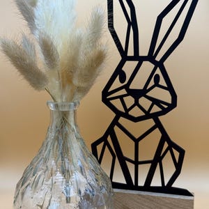 Peut inclure: Une sculpture de lapin géométrique noire sur une base en bois. Le lapin est fait de métal noir fin et a un design géométrique. Le lapin est debout sur une base en bois. Le lapin est devant un vase avec de l'herbe de pampa séchée.