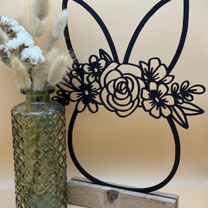Könnte beinhalten: Schwarze Metall-Hasensilhouette mit einem Blumenkranz um den Kopf. Der Hase steht auf einem Holzsockel.