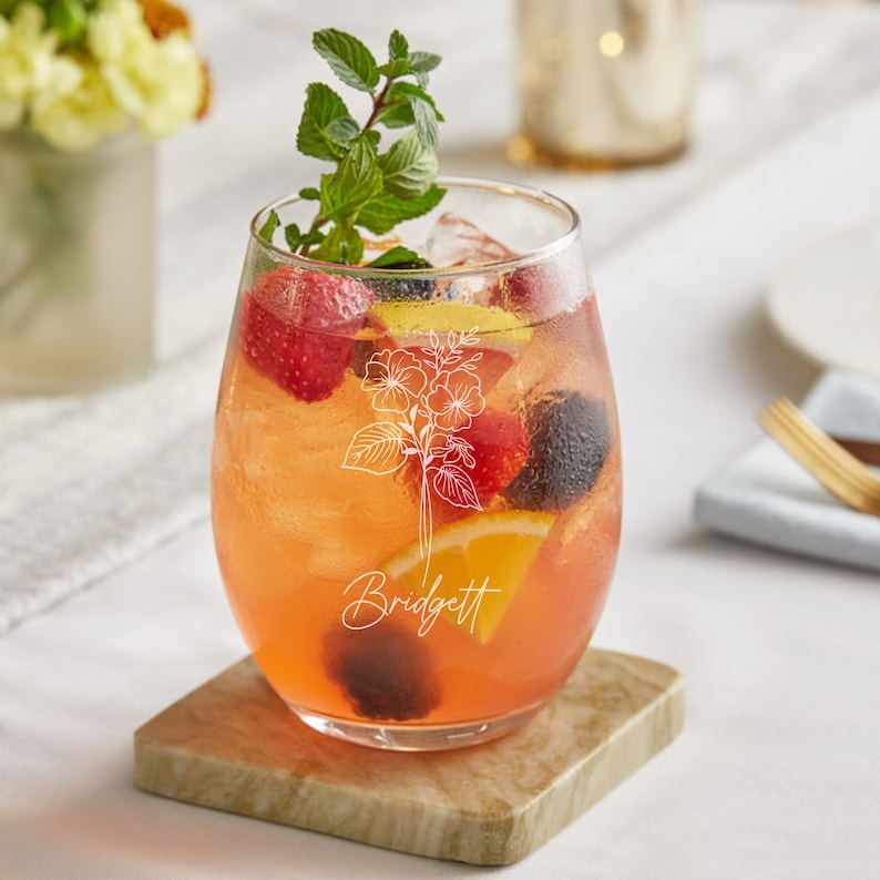May include: A clear glass with a stem, filled with a pink drink and fruit, including strawberries, blackberries, and orange slices. The glass has a floral design and the name "Bridgett" etched on the side.