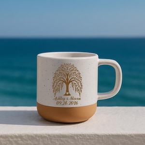 May include: A white ceramic mug with a brown base, featuring a gold tree design and the names "Ashley & Steven" with the date "09.26.2016". The mug has a large handle and is set against a blue ocean backdrop.