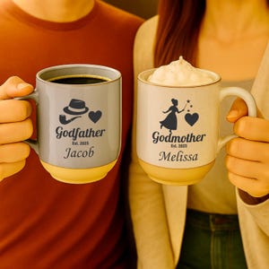 May include: Two ceramic mugs with a two-tone design. The left mug is gray with a fedora, pipe, heart, and the text "Godfather Est. 2025 Jacob". The right mug is cream with a fairy, heart, and the text "Godmother Est. 2025 Melissa" filled with foam.