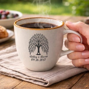 May include: A cream-colored coffee mug with a brown rim, filled with dark coffee and steam rising. The mug features a black tree design with the names "Ashley & Steven" and the date "09.26.2017" printed below the tree.
