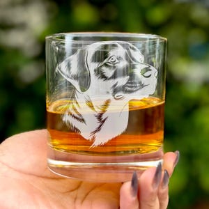 May include: A clear glass tumbler with a black and white etched image of a dog's head. The glass is filled with a golden brown liquid.
