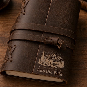 May include: A dark brown leather journal with a wrap-around strap closure. The cover features an engraved graphic of a bear in a mountain forest landscape with the text Into the Wild. The binding includes decorative leather lacing on the side.