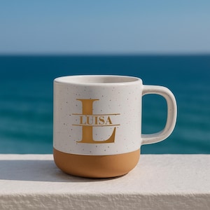 May include: A speckled white ceramic mug with a brown base and a large gold letter "L" with the name "LUISA" printed below. The mug has a curved handle and is set against a backdrop of the ocean.