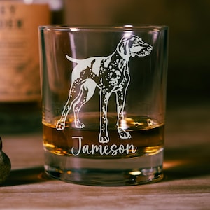 May include: A clear rocks glass with a white etched illustration of a dog. The dog has a spotted coat and is standing. The word "Jameson" is written in cursive at the bottom of the glass. The glass is filled with a golden liquid.