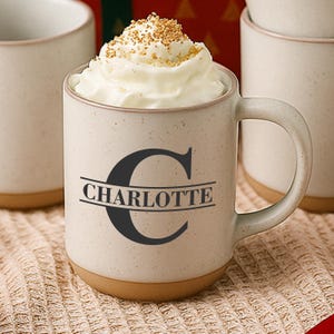 May include: A speckled off-white ceramic mug with a handle, featuring a large black letter C and the name CHARLOTTE printed in black across the center. The mug is filled with whipped cream topped with brown sprinkles, and has a tan base.