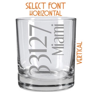 May include: A clear glass tumbler with the text "33127 Miami" etched on the side in a grey font. The text is oriented vertically. The tumbler is on a white background.