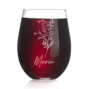 May include: A clear glass wine glass with a red wine inside. The glass has an etched design of a lily of the valley flower and the name "Maria" on the bottom.