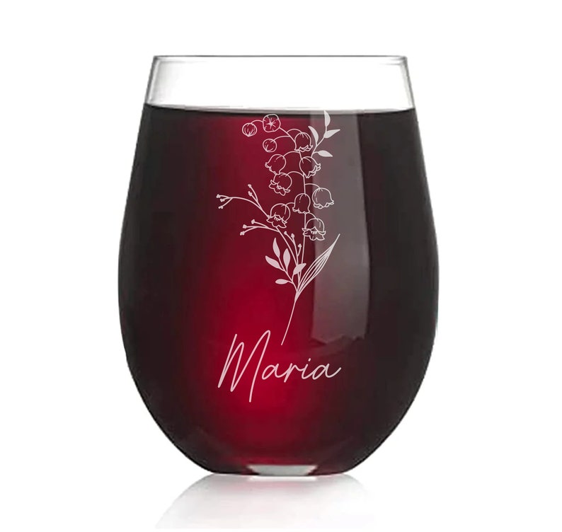 May include: A clear glass wine glass with a red wine inside. The glass has an etched design of a lily of the valley flower and the name "Maria" on the bottom.