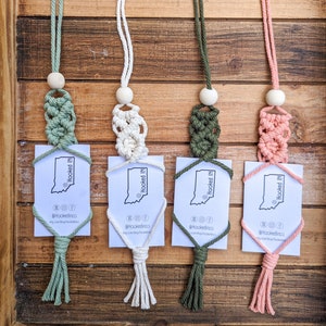 May include: Four macrame keychains with tassels in green, white, and pink. Each keychain features a small wooden bead and a business card with the text "Hooked IN" and the Instagram handle "@hookedinco".