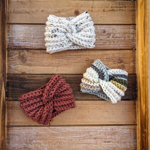 May include: Three crocheted headbands featuring a twisted front design. One headband is solid cream with dark flecks, one is solid dark rust, and one features horizontal stripes in cream, sage green, dark brown, and light gray yarn textures.