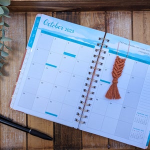 May include: An open planner with a blue and white watercolor design. The planner is open to October 2023 and has a brown tassel bookmark with a quote from Walt Disney. "All our dreams can come true, if we have the courage to pursue them." - Walt Disney