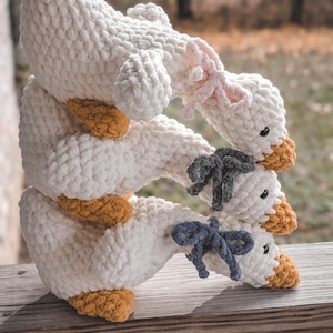 May include: Three stacked, handmade crochet geese. Each goose is white with a yellow-orange beak and feet. The top goose has a pink bow, the middle goose has a gray bow, and the bottom goose has a blue bow. The geese are on a wooden surface.