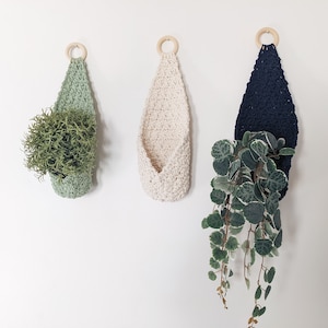 May include: Three crochet plant holders in light green, cream, and navy blue hang on a white wall. Each holder has a wooden ring at the top for hanging. The light green holder contains a green plant, while the navy blue holder holds a trailing plant.