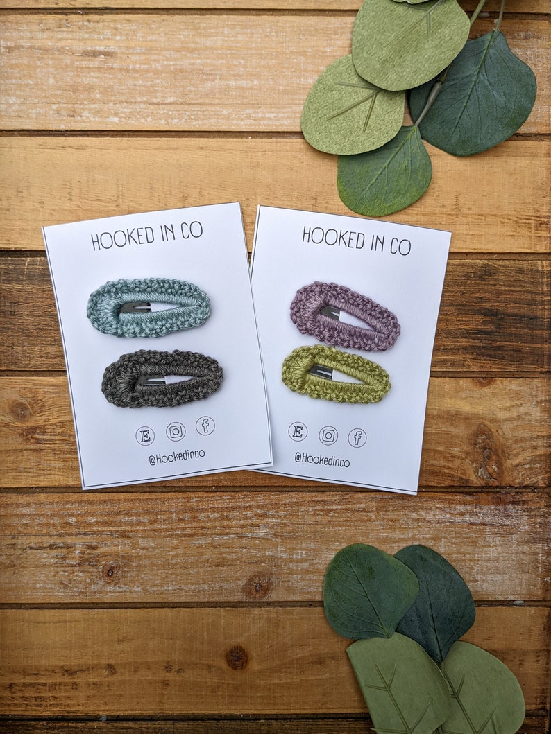 Puede incluir: Dos juegos de tres pinzas para el pelo de crochet en tarjetas blancas. Las tarjetas tienen el texto "HOOKED IN CO" y el nombre de usuario de las redes sociales "@hookedinco". Las pinzas son en tonos de verde, azul y morado.