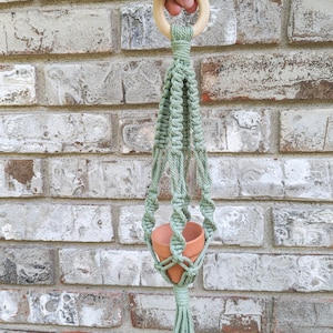 May include: A light green macrame plant hanger with a wooden ring at the top and a terracotta pot hanging in the middle.
