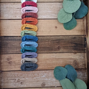 Puede incluir: Una colecci&oacute;n de 14 pinzas para el pelo de crochet en varios colores, incluyendo blanco, rojo, rosa, marr&oacute;n, amarillo, verde, azul, morado y gris. Las pinzas est&aacute;n dispuestas verticalmente sobre una superficie de madera.