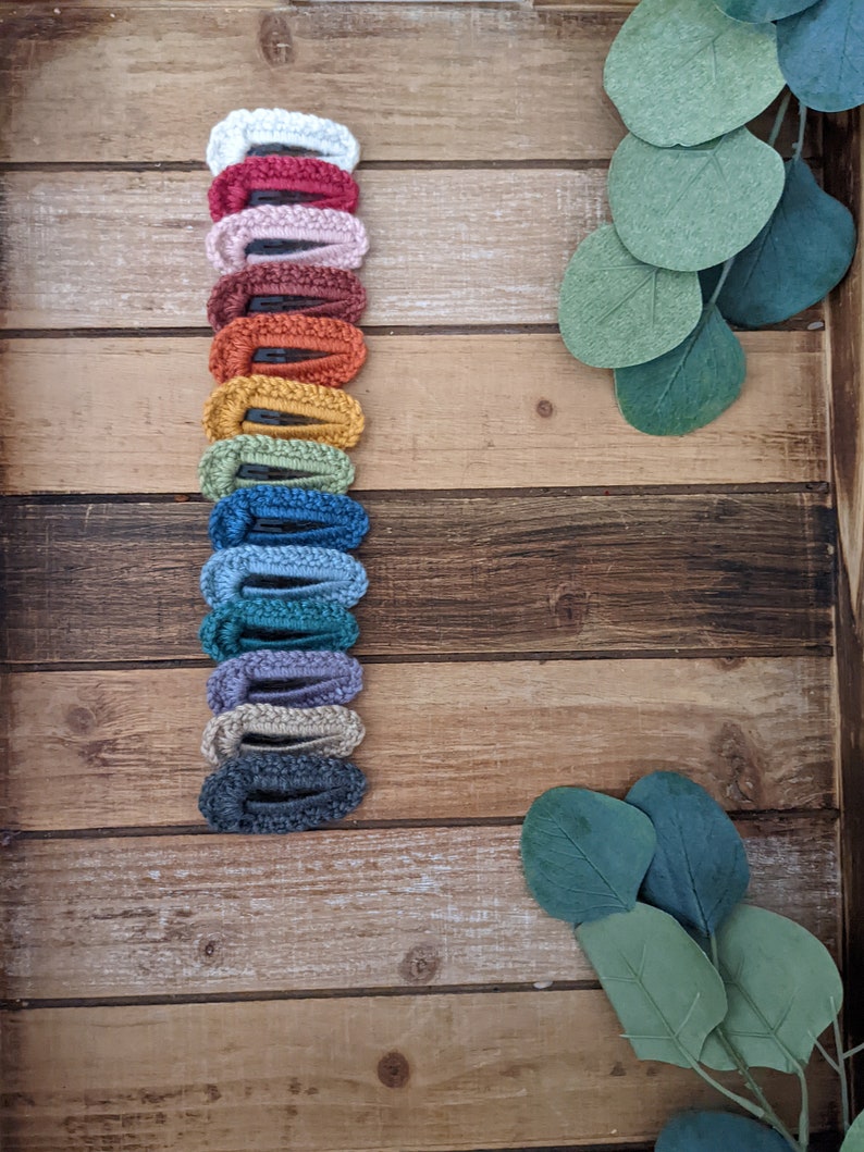 Puede incluir: Una colecci&oacute;n de 14 pinzas para el pelo de crochet en varios colores, incluyendo blanco, rojo, rosa, marr&oacute;n, amarillo, verde, azul, morado y gris. Las pinzas est&aacute;n dispuestas verticalmente sobre una superficie de madera.